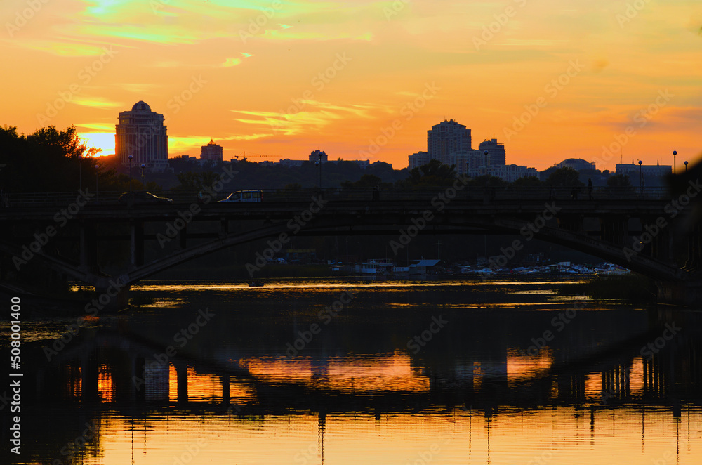 Magic sunset in Kyiv. Silhouettes of buildings and a bridge reflected in tranquil water of Rusanivka's channel. Vibrant orange sunset colors