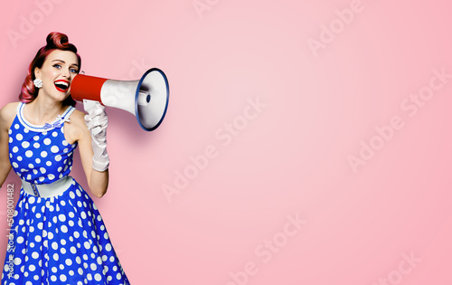 Portrait image of purple haired woman holding mega phone, shout advertising something. Girl in blue pin up style dress with mega phone loudspeaker. Isolated on rose pink background. Beauty model.