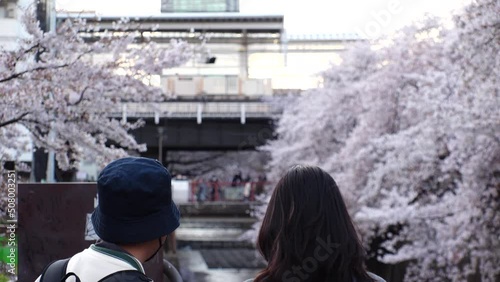 NAKAMEGURO, TOKYO, JAPAN - APR 2022 : Cherry blossoms at Meguro river and unidentified people wearing masks to protect from Coronavirus. Japanese spring season in COVID-19 pandemic. Slow motion shot. photo