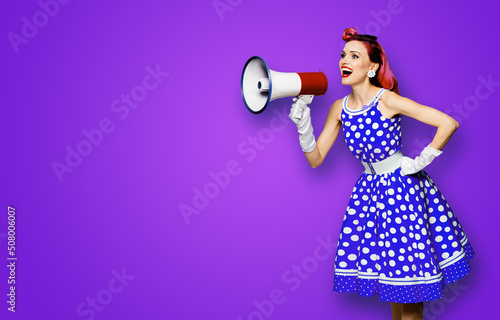 Purple haired woman holding red megaphone, shout advertising something. Girl in blue pin up style with mega phone loudspeaker. Violet background with mock up. Female model in retro fashion dress.