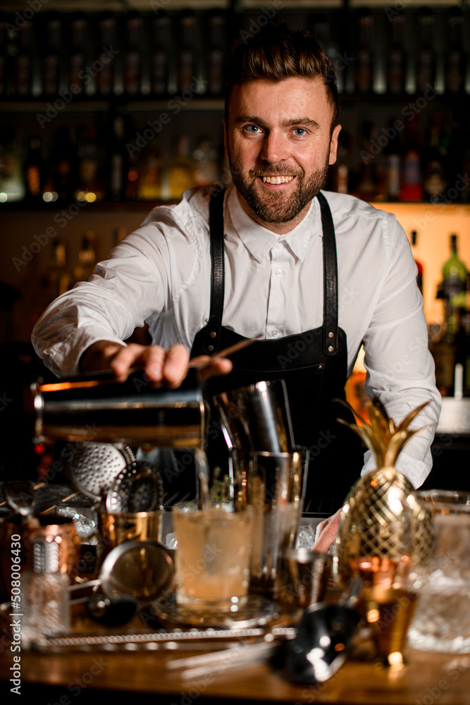 focus on smiling bartender pouring a drink from a steel shaker cup into crystal glass focus on smiling bartender pouring a drink from a steel shaker cup into crystal glass