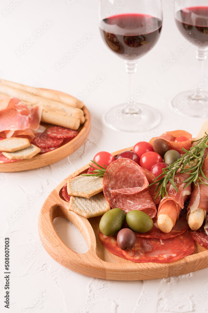 Charcuterie plate with different types of meat snacks - proscuitto. Wooden plates with traditional italian antipasti served with glasses of red wine on white concrete table. Vertical image