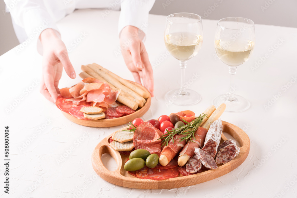 Woman serving traditional Italian antipasti on white table. Charcuterie plate with different types of sausages and glasses of white wine - salami, bresaola, proscuitto served with olives and grissini