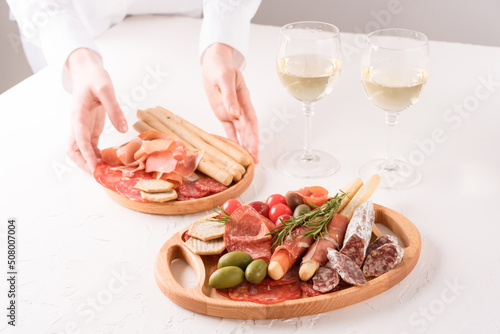 Woman serving traditional Italian antipasti on white table. Charcuterie plate with different types of sausages and glasses of white wine - salami, bresaola, proscuitto served with olives and grissini