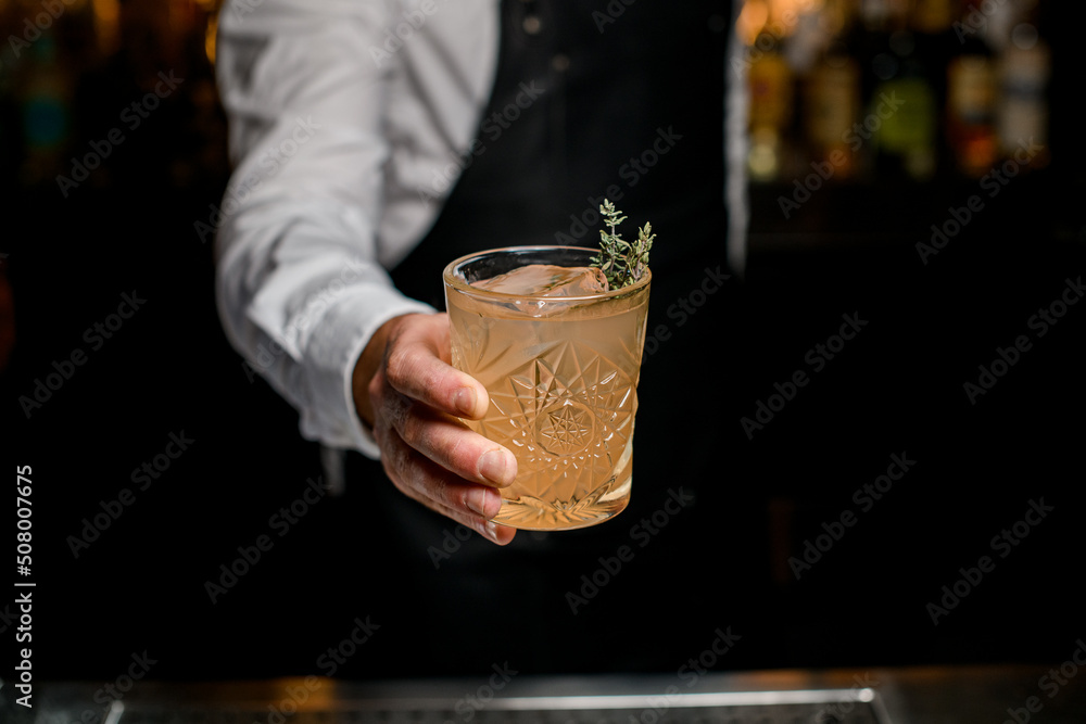 great view of glass with cocktail garnished with rosemary in the hand of bartender on blurred background