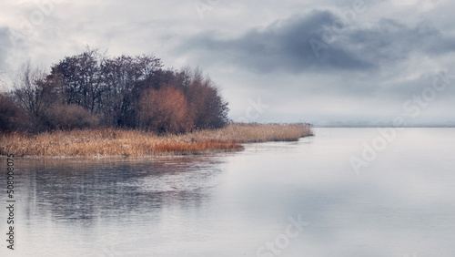 Fototapeta Naklejka Na Ścianę i Meble -  Autumn landscape with an island on a lake or river in cloudy weather, reflection of trees and clouds in the river water. Gloomy autumn morning