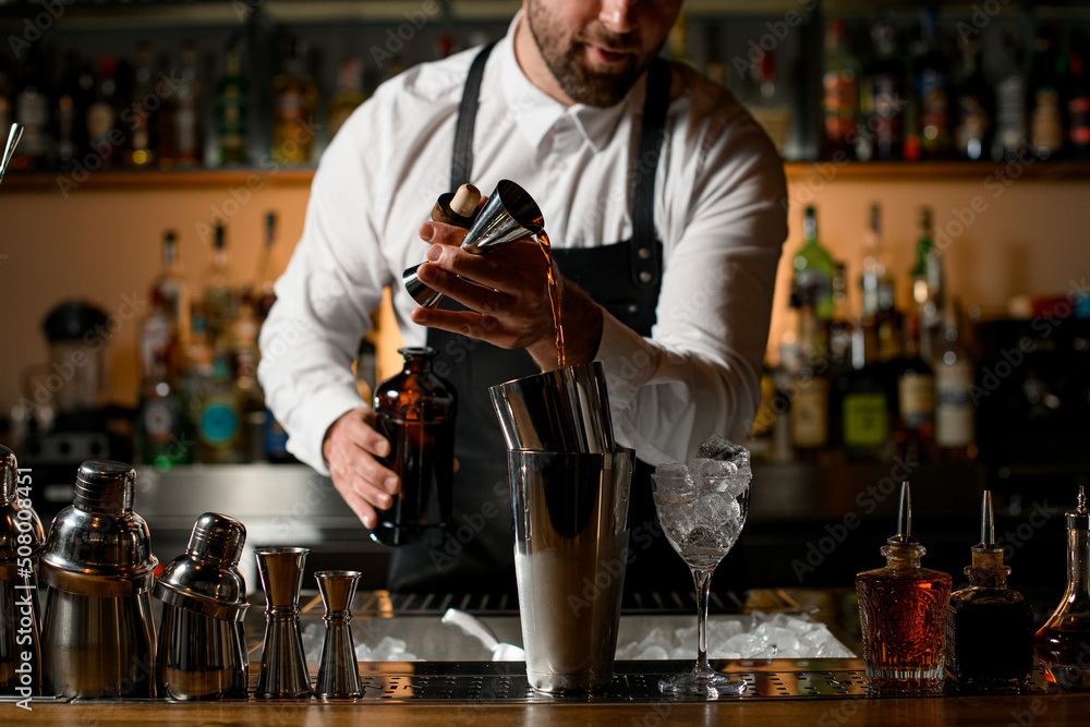 different steel shakers and bottles stand on the bar counter, and hand of bartender pours alcoholic drink into cup