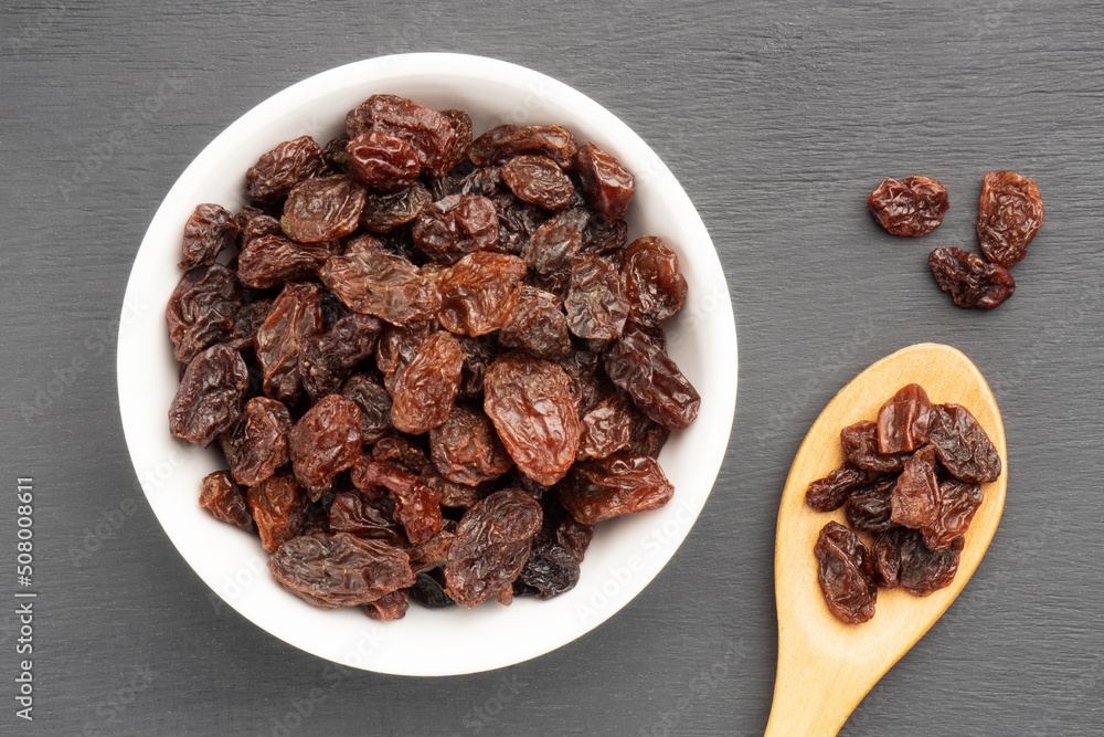 Raisin in white ceramic bowl and wooden spoon on black wooden background. Macro. Vegetarian concept Raisin in white ceramic bowl and wooden spoon on black wooden background. Macro. Vegetarian concept