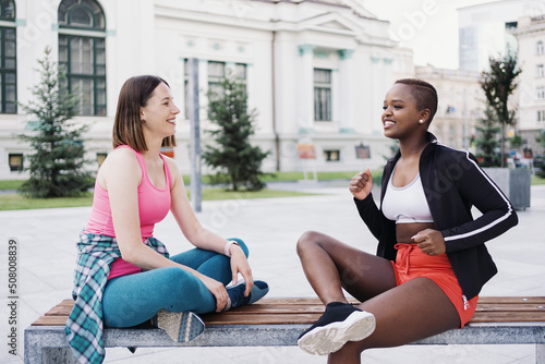 Cheerful smiling friends in sportswear sitting on bench in the city dicussing in park. Multiethnic women having a fitness workout break.