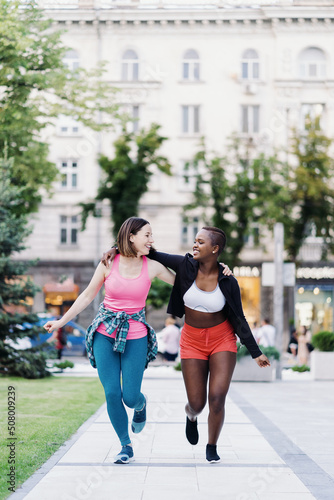 Cheerful smiling friends in sportswear running in the city dicussing. Multiethnic women having a fitness workout.