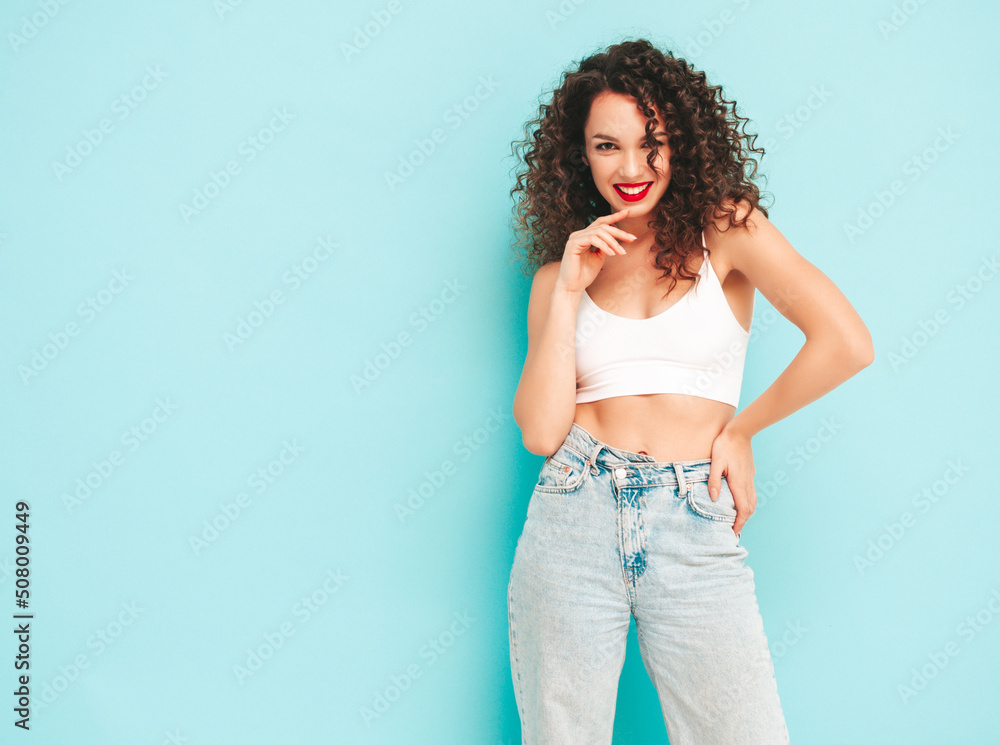 Sexy young beautiful smiling female in trendy summer hipster clothes. carefree woman posing near blue wall in studio. Positive brunette model with afro curls hairstyle. Cheerful and happy Sexy young beautiful smiling female in trendy summer hipster clothes. carefree woman posing near blue wall in studio. Positive brunette model with afro curls hairstyle. Cheerful and happy