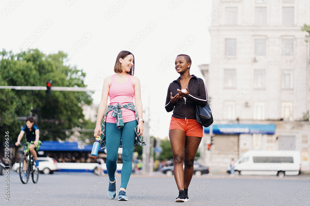 Cheerful smiling friends in sportswear walking after a sport session in the city discussing and having fun. Multiethnic women having a fitness workout jogging.