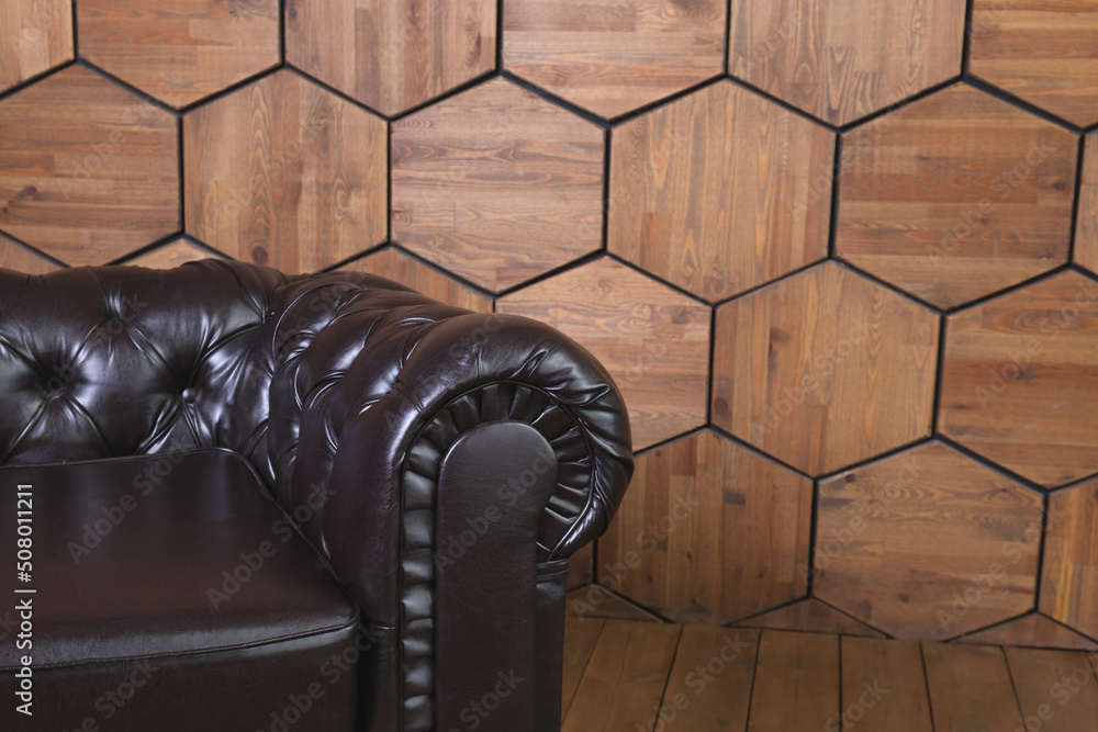 Low black leather sofa with a round armrest set on hexagon wood-panelled wall background. An empty leather couch with a low back. Space for text. Low black leather sofa with a round armrest set on hexagon wood-panelled wall background. An empty leather couch with a low back. Space for text.