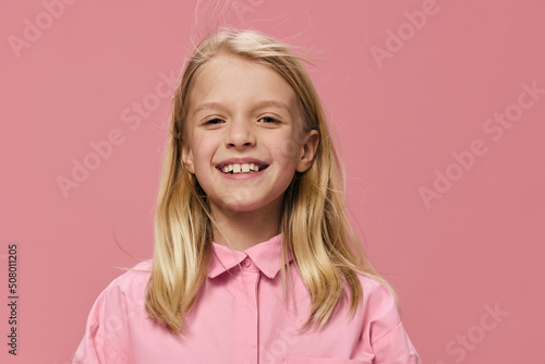 horizontal portrait of a school-age blonde girl in a pink shirt, standing against a one-time background and smiling broadly looking at the camera. Studio photo with empty space for advertising mockup