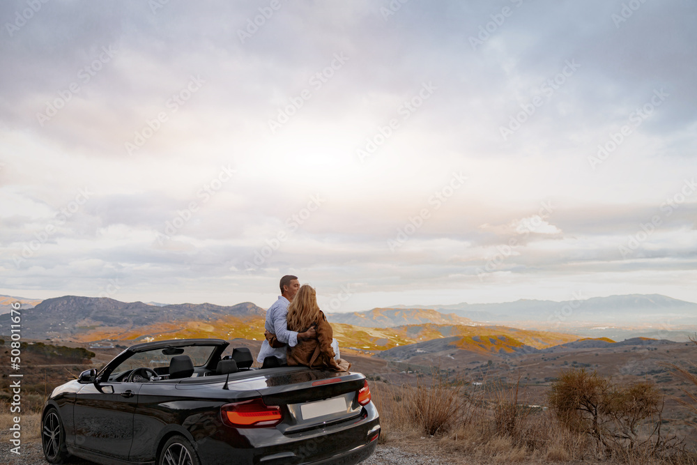 Rear of cute couple standing by convertible car cabriolet looking at panorama view. Romantic concept