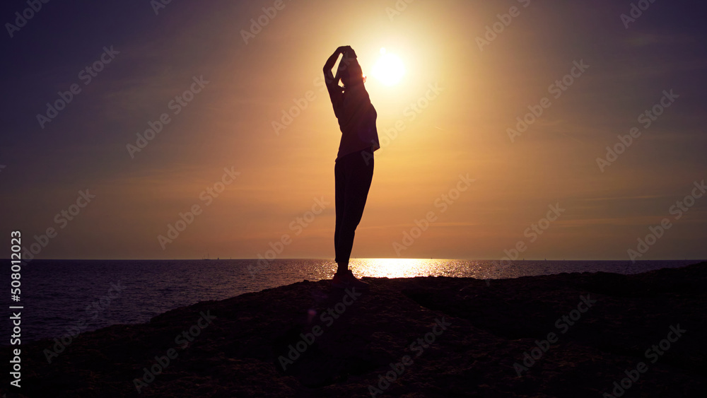 Silhouette of a woman stretching and relaxing while watching sunset sunrise above the ocean.