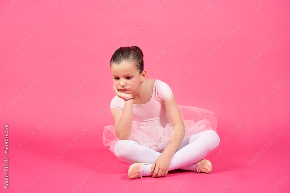 Bored little ballet dancer girl sitting on the floor. Studio shot with pink background Bored little ballet dancer girl sitting on the floor. Studio shot with pink background