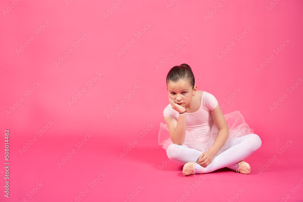Bored little ballet dancer girl sitting on the floor and looking at camera. Studio shot with vivid pink background.