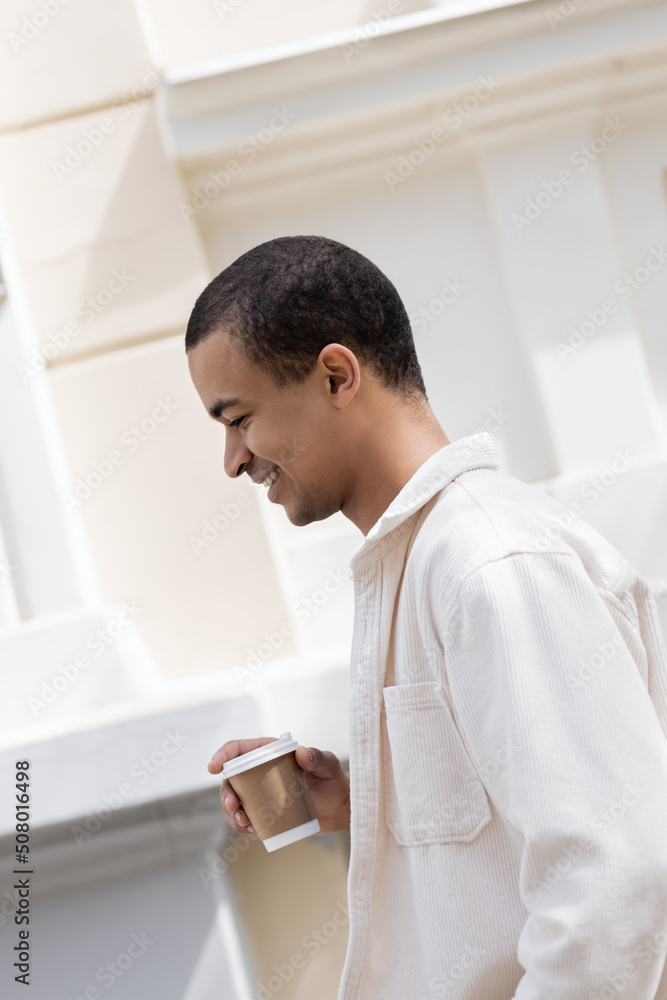 side view of happy african american man in shirt jacket holding coffee to go and walking in urban city.
