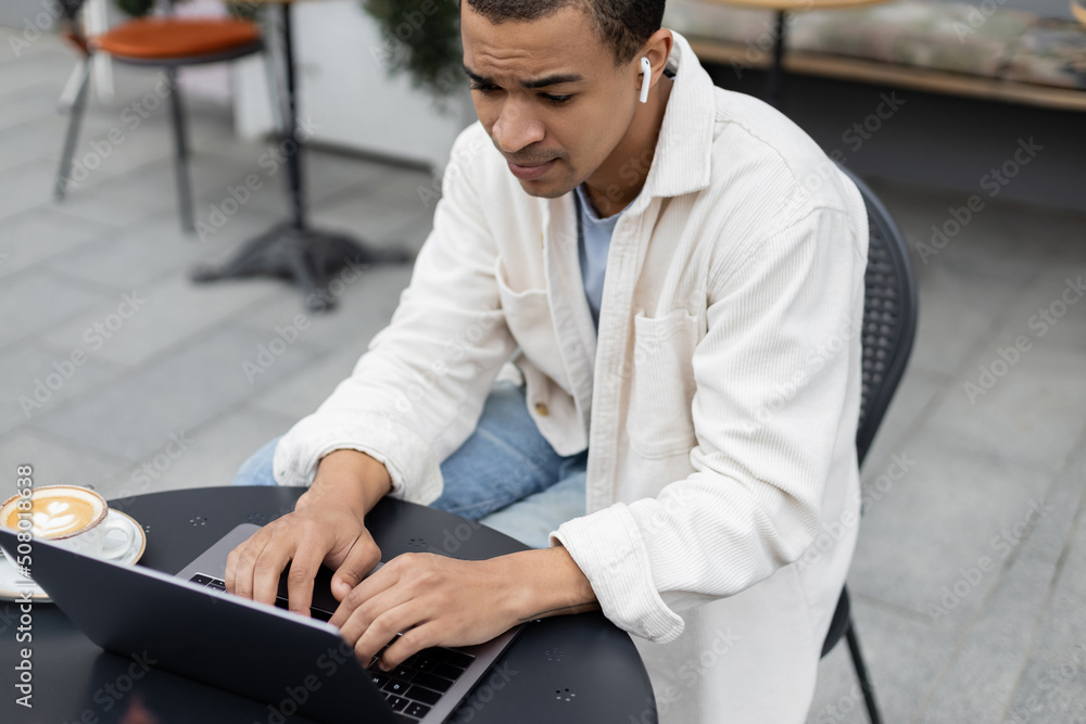 african american freelancer in wireless earphones using laptop near cappuccino on summer terrace. african american freelancer in wireless earphones using laptop near cappuccino on summer terrace.