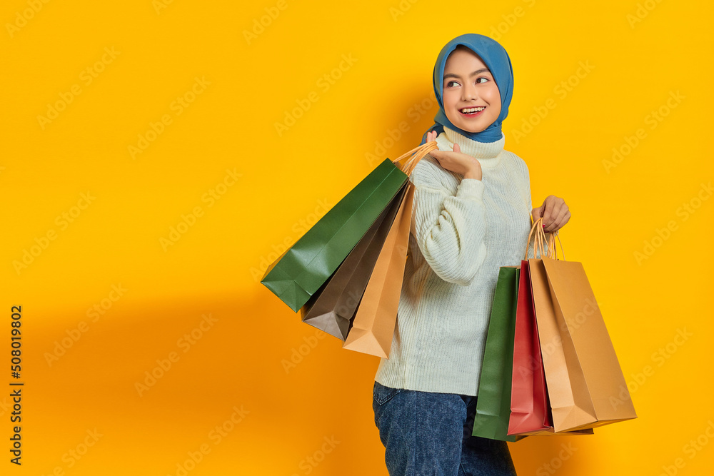 Cheerful beautiful Asian woman in white sweater showing so many shopping bags, shopping discount promo isolated over yellow background