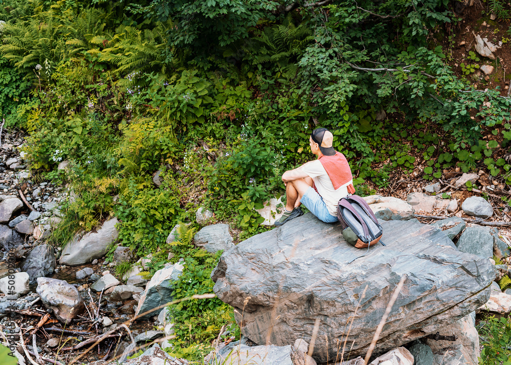 Fototapeta premium Young man in a cap with a backpack sitting on a large stone and looking at a mountain stream among the forest and lush vegetation in a summer hike Enjoying nature Active lifestyle