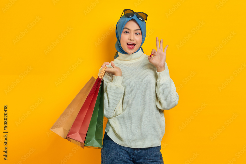 Cheerful beautiful Asian woman in white sweater holding shopping bags, showing okay gesture isolated over yellow background