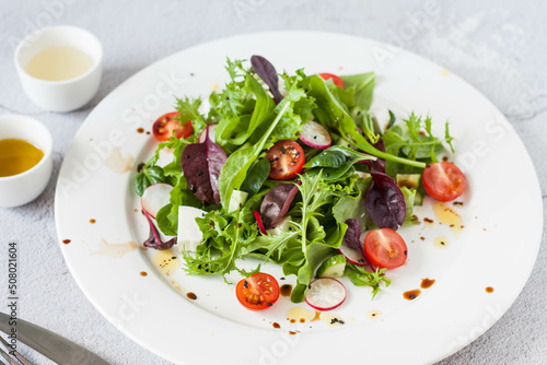 Green salad of fresh lettuce leaves, cherry tomatoes, feta cheese and assorted seeds with a sauce of honey, olive oil and balsamic vinegar on a flat plate on a stone table. Healthy vegan food.