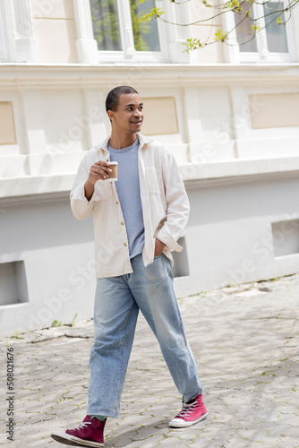 full length of positive african american man in shirt jacket and jeans holding coffee to go and walking in urban city.