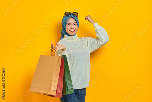Cheerful beautiful Asian woman in white sweater holding shopping bags and celebrate luck isolated over yellow background