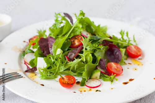 Green salad of fresh lettuce leaves, cherry tomatoes, feta cheese and assorted seeds with a sauce of honey, olive oil and balsamic vinegar on a flat plate on a stone table. Healthy vegan food.