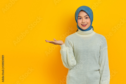 Cheerful beautiful Asian woman in white sweater presenting copy space on palm isolated over yellow background © Bangun Stock Photo