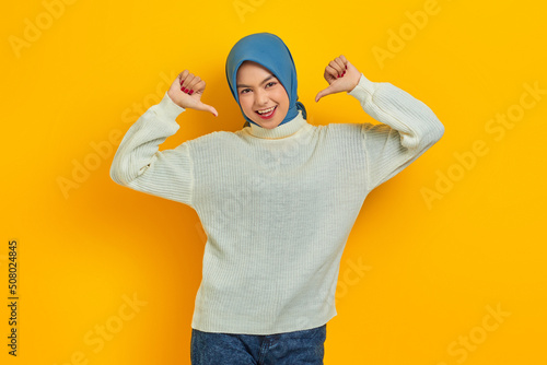 Overjoyed beautiful Asian woman in white sweater pointing thumbs at herself with proud isolated over yellow background photo