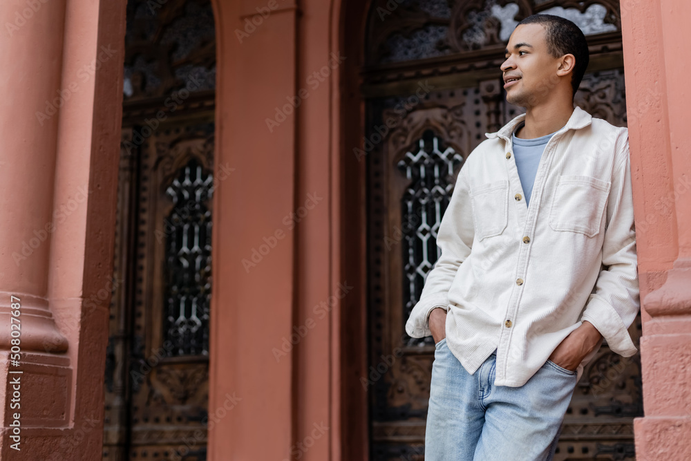 joyful african american man in shirt jacket posing with hands in pockets near building in europe. joyful african american man in shirt jacket posing with hands in pockets near building in europe.