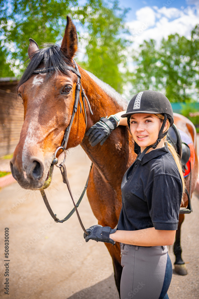 Fototapeta premium Lovely young woman wearing helmet stroking to her brown horse