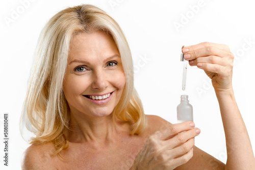 Sophisticated middle aged woman applies regenerating serum on the face. Charming blonde 40s lady using anti-aging complex with hyaluronic acid isolated on white background. Skincare concept