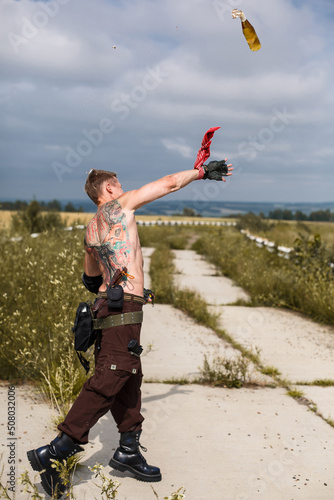 male vandal with dirty naked torso throws Molotov cocktail, against backdrop of an overgrown abandoned road
