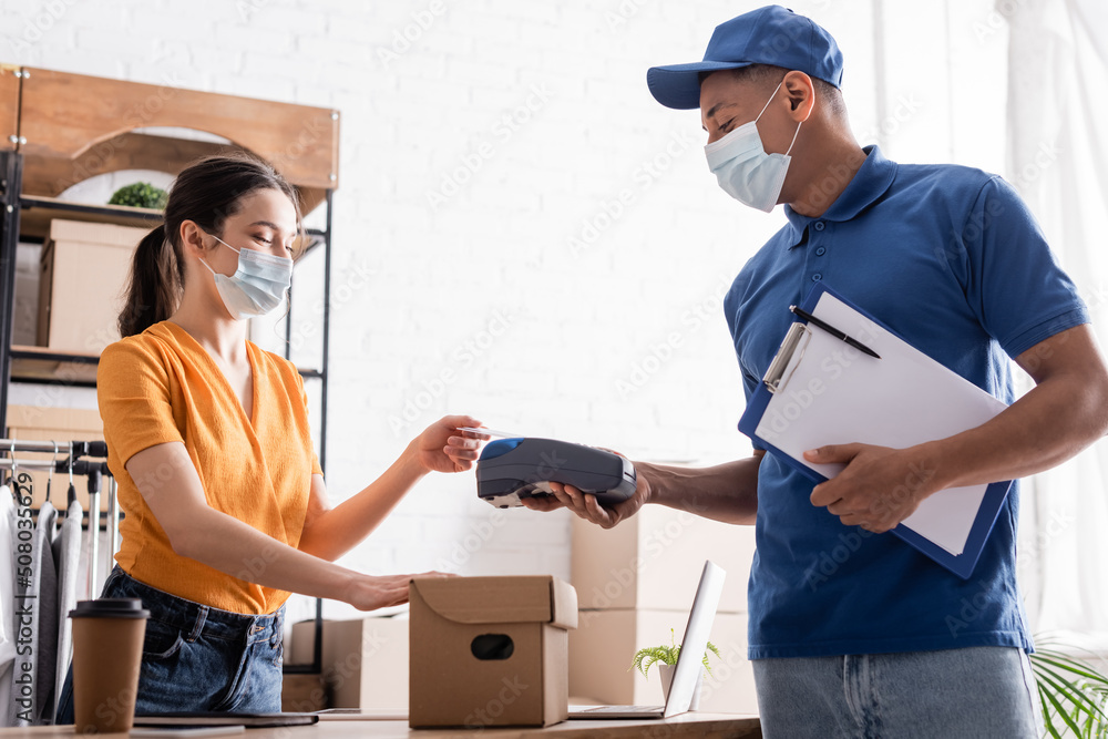 Fototapeta premium Seller in medical mask paying with credit card near african american courier and carton box in online web store.
