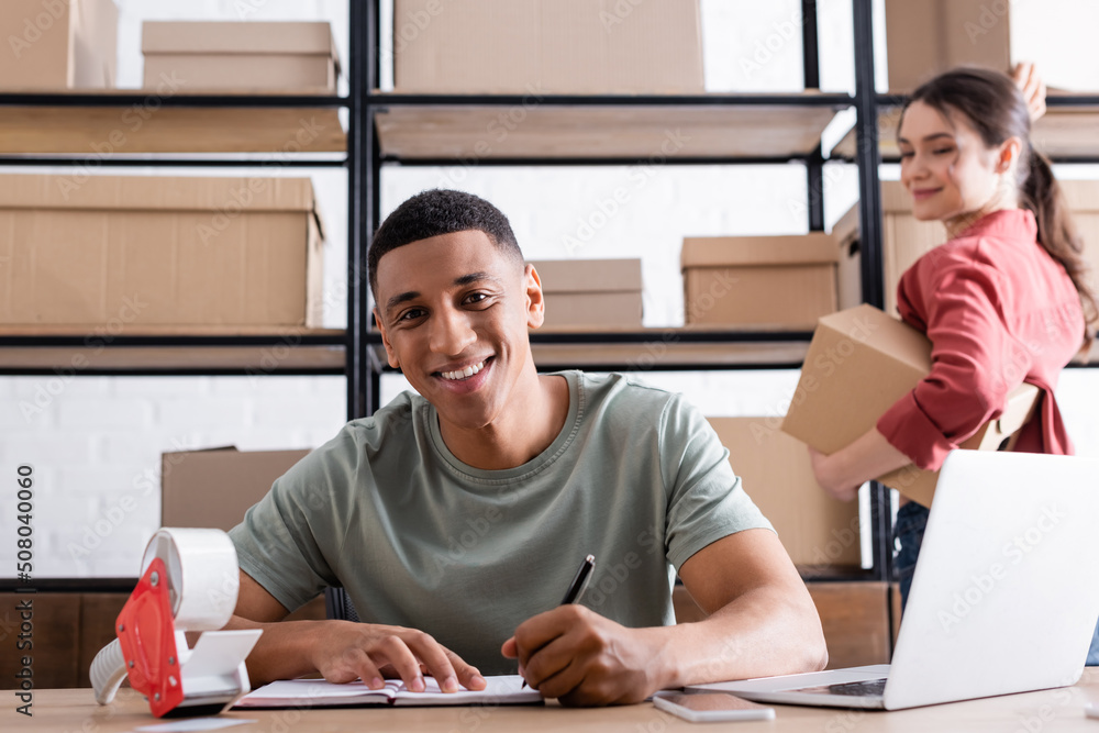 Smiling african american online web store owner writing on notebook near blurred colleague with boxes. Smiling african american online web store owner writing on notebook near blurred colleague with boxes.