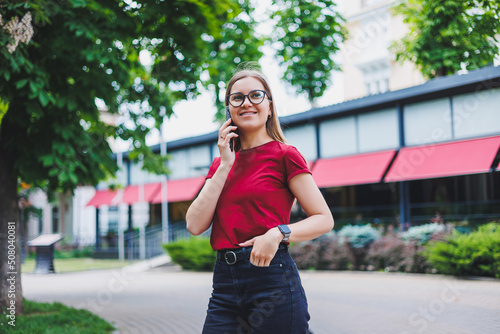 Young cheerful female in grey pullover and blue jeans and glasses standing on sidewalk carrying shopping bags and messaging on smartphone in New York © Дмитрий Ткачук