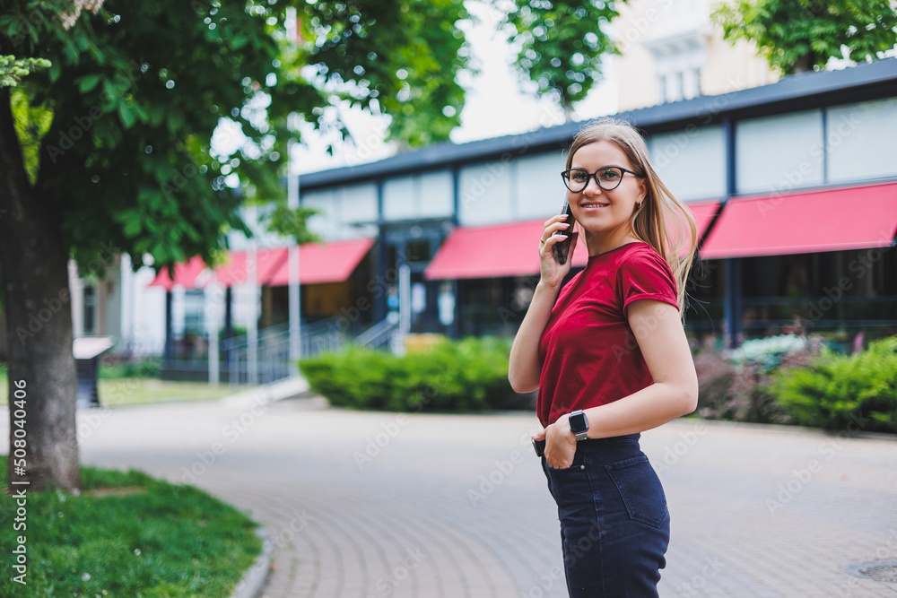 Young cheerful female in grey pullover and blue jeans and glasses standing on sidewalk carrying shopping bags and messaging on smartphone in New York Young cheerful female in grey pullover and blue jeans and glasses standing on sidewalk carrying shopping bags and messaging on smartphone in New York