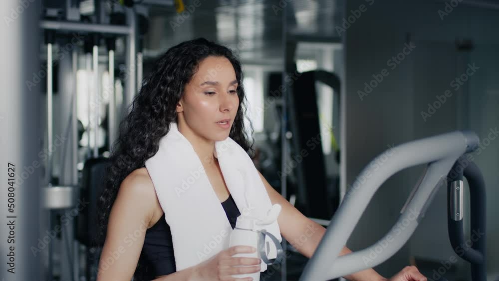 Water balance during sport training. Young active lady drinking from bottle, exercising on training apparatus at gym Water balance during sport training. Young active lady drinking from bottle, exercising on training apparatus at gym