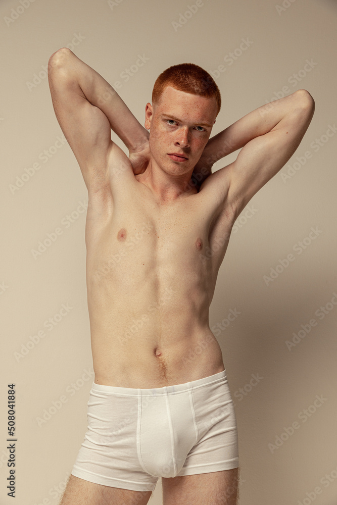 Half-length portrait of young muscular red-haired man posing in white underwear, boxers isolated over grey studio background. Serious look