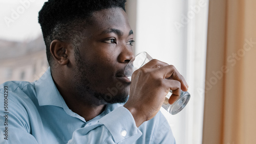 African american young dreaming man thoughtful positive guy turn head look at window drink fresh cold water glass of pure liquid hydrate thirst suffer from hot make pause feeling rejuvenation energy