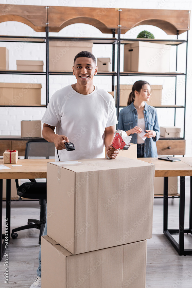 Positive african american online web store owner holding scanner and adhesive tape near blurred colleague. Positive african american online web store owner holding scanner and adhesive tape near blurred colleague.