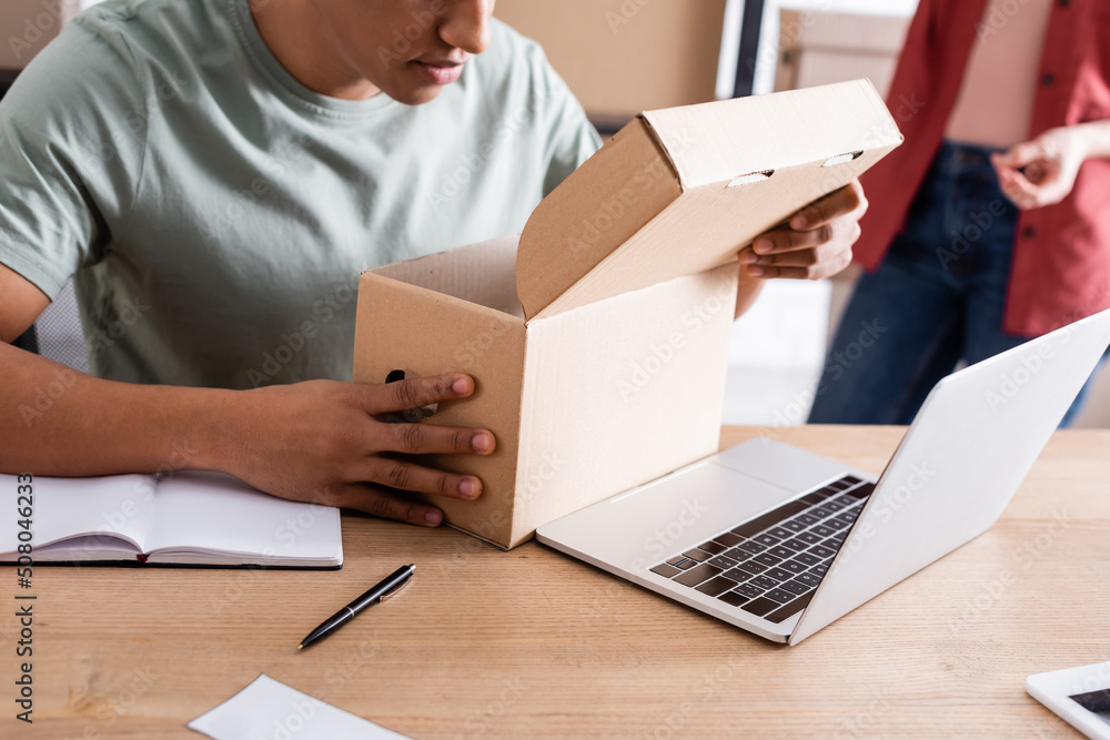 Cropped view of african american seller holding carton box near laptop in online web store. Cropped view of african american seller holding carton box near laptop in online web store.