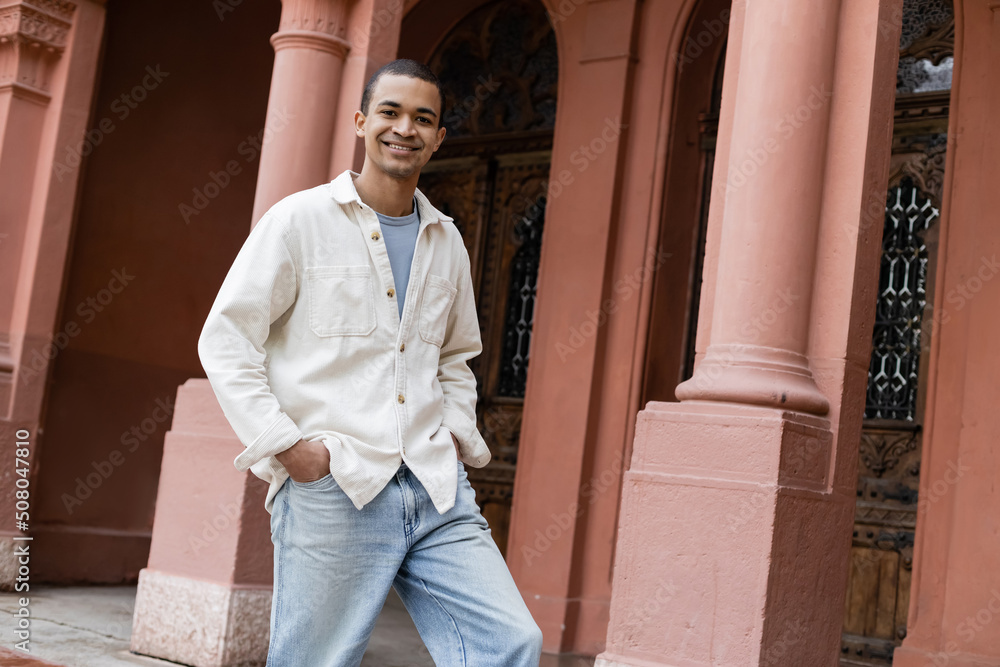 joyful african american man in shirt jacket standing with hands in pockets near building.