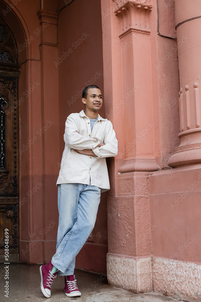 full length of smiling african american man in shirt jacket posing with crossed arms near building in europe. full length of smiling african american man in shirt jacket posing with crossed arms near building in europe.