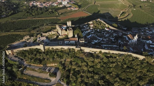 Ancient castle fortress in Portugal drone flyby with green fields on sunny day. Old European middle ages town Obidos with modern bridge highway in the background.