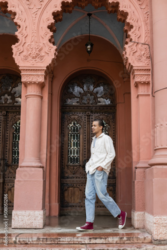 full length of happy african american man in shirt jacket walking with hands in pockets near building in europe.